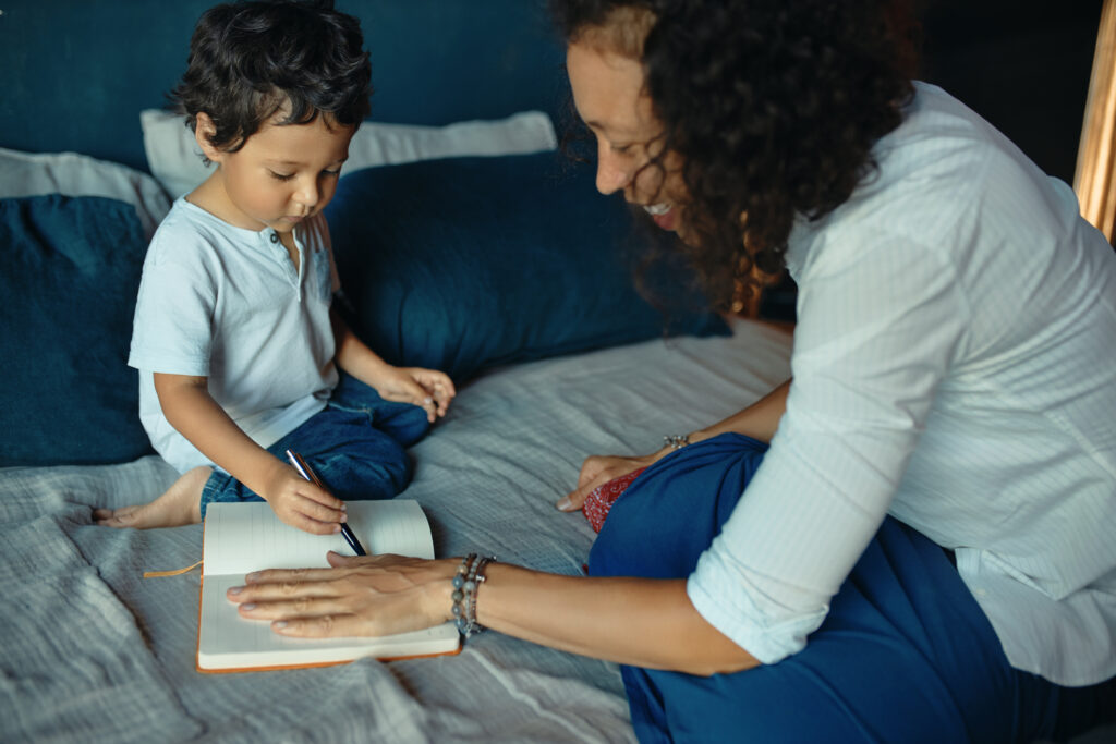 Young Latin woman and her son tracing outlines in a notebook on a bed, emphasizing family support and nurturing in the context of child custody and financial well-being.