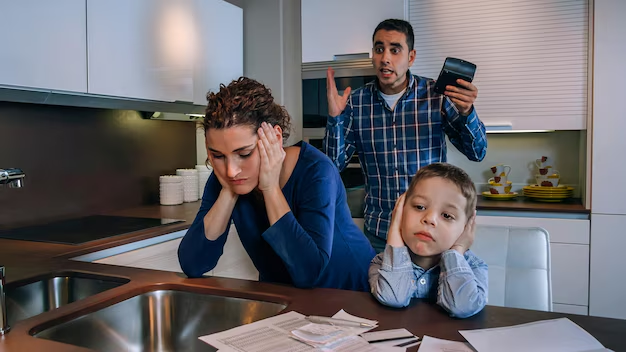 Frustrated family in kitchen dealing with financial stress, woman holding head in hands, child covering ears, man gesturing with phone, highlighting family law challenges.