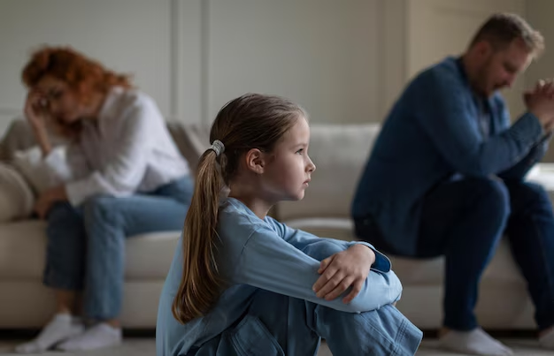 Child sitting on the floor, looking concerned, with parents in the background appearing distressed, reflecting family conflict and the emotional impact of divorce or custody issues.
