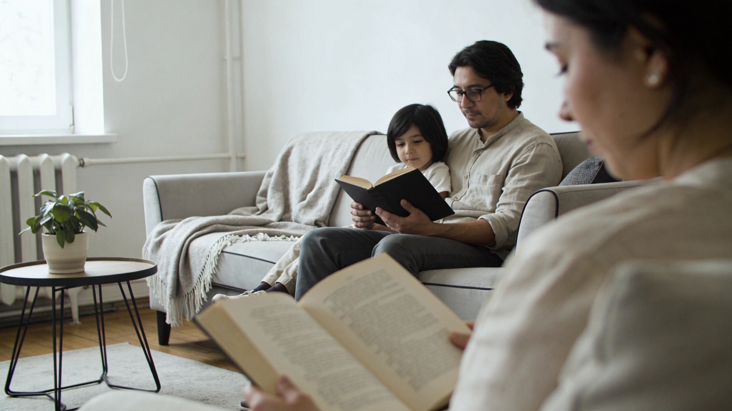 A father reading a book to his young son while the mother reads nearby in a living room.